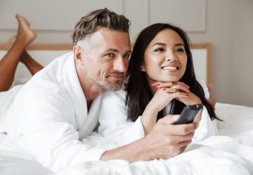 Photo of multiethnic couple man and woman wearing white bathrobe lying together in home bedroom or hotel apartment with remote control while watching tv