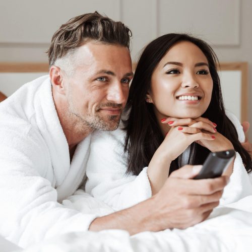 Photo of multiethnic couple man and woman wearing white bathrobe lying together in home bedroom or hotel apartment with remote control while watching tv